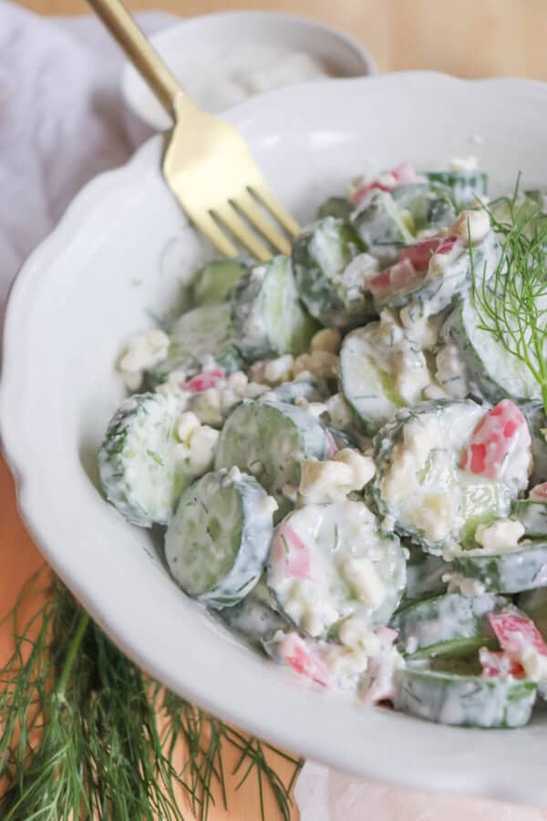 Sliced cucumbers in tzatziki with diced pickled onions and dill in a white bowl with a gold fork in it.