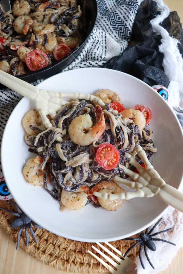 Purple and white linguine noodles in a cream sauce topped with shrimp and cherry tomatoes with skeleton hands in a white bowl.