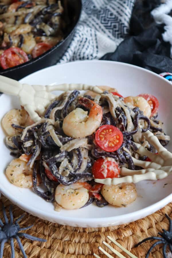 Purple and white linguine noodles in a cream sauce topped with shrimp and cherry tomatoes with skeleton hands in a white bowl.