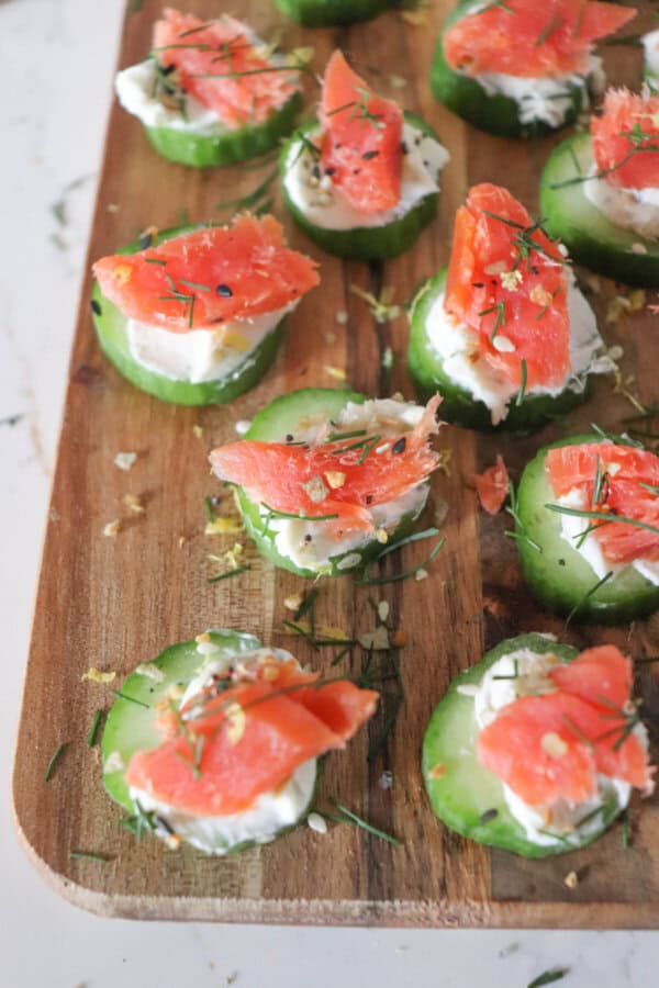Small rounds of cucumber slices topped with cream cheese, smoked salmon and garnished with fresh dill and lemon zest on a wooden cutting board.