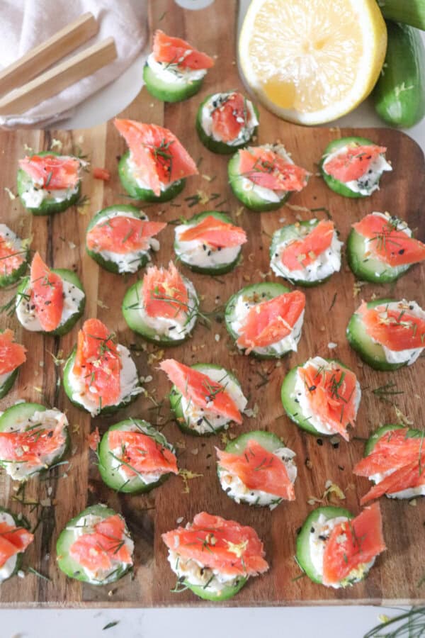 Small rounds of cucumber slices topped with cream cheese, smoked salmon and garnished with fresh dill and lemon zest on a wooden cutting board.