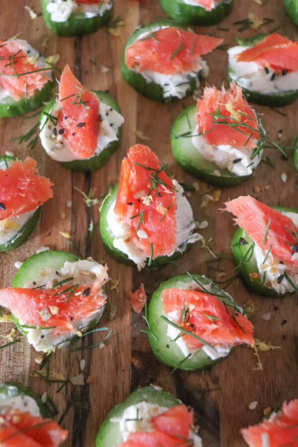 Small rounds of cucumber slices topped with cream cheese, smoked salmon and garnished with fresh dill and lemon zest on a wooden cutting board.