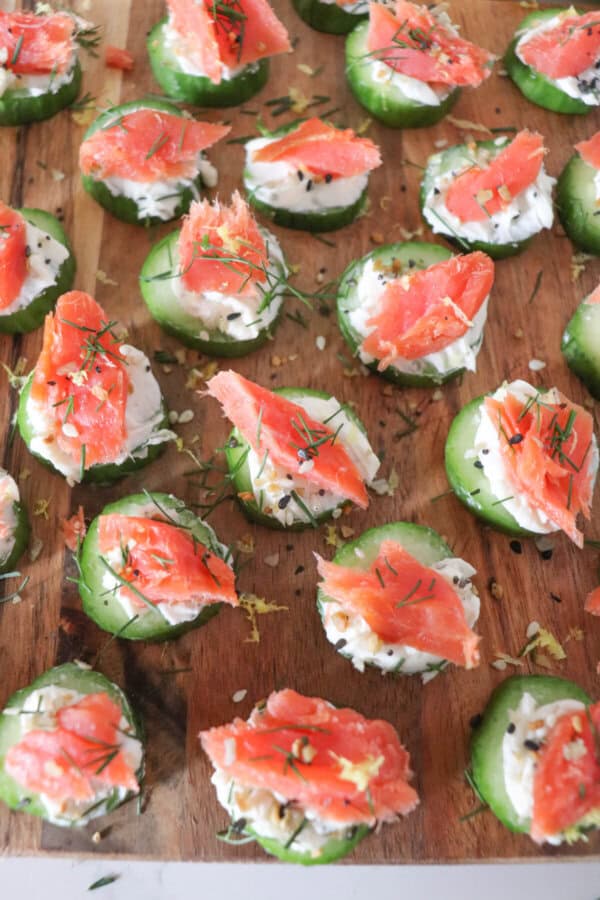 Small round of cucumber slices topped with cream cheese, smoked salmon and garnished with fresh dill and lemon zest on a wooden cutting board.