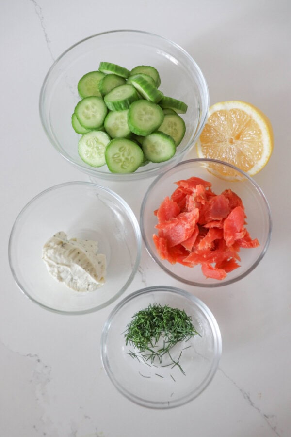 Ingredients for smoked salmon cucumber bites in clear glass bowls on a white counter.