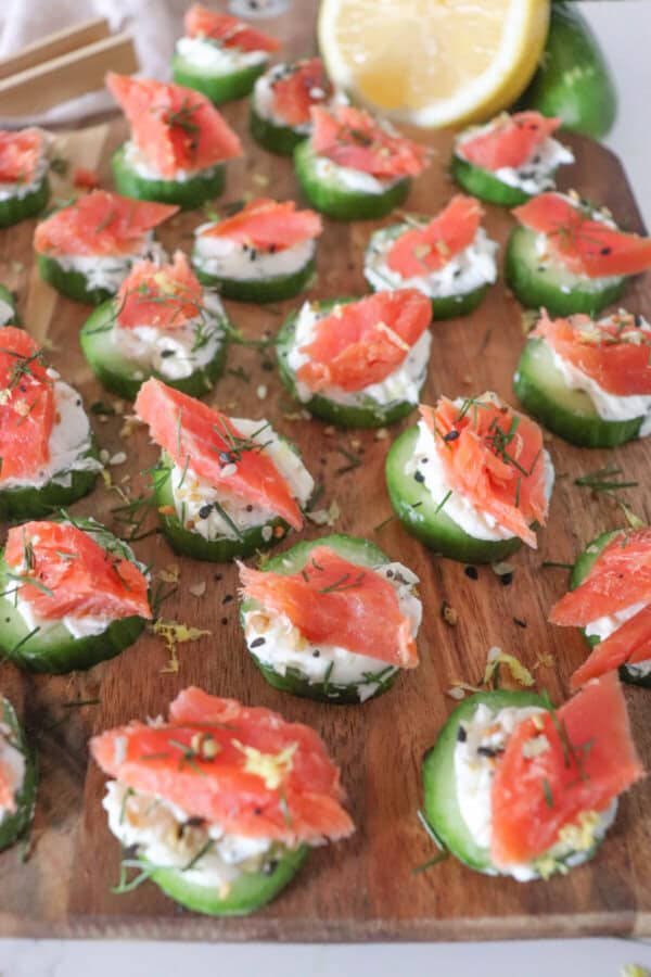 Small rounds of cucumber slices topped with cream cheese, smoked salmon and garnished with fresh dill and lemon zest on a wooden cutting board.
