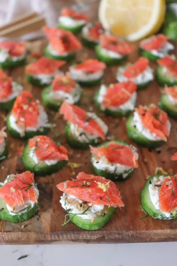 Small rounds of cucumber slices topped with cream cheese, smoked salmon and garnished with fresh dill and lemon zest on a wooden cutting board.