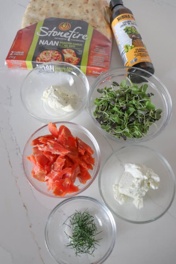 Ingredients for smoked salmon flatbread in clear glass bowls on a white surface.