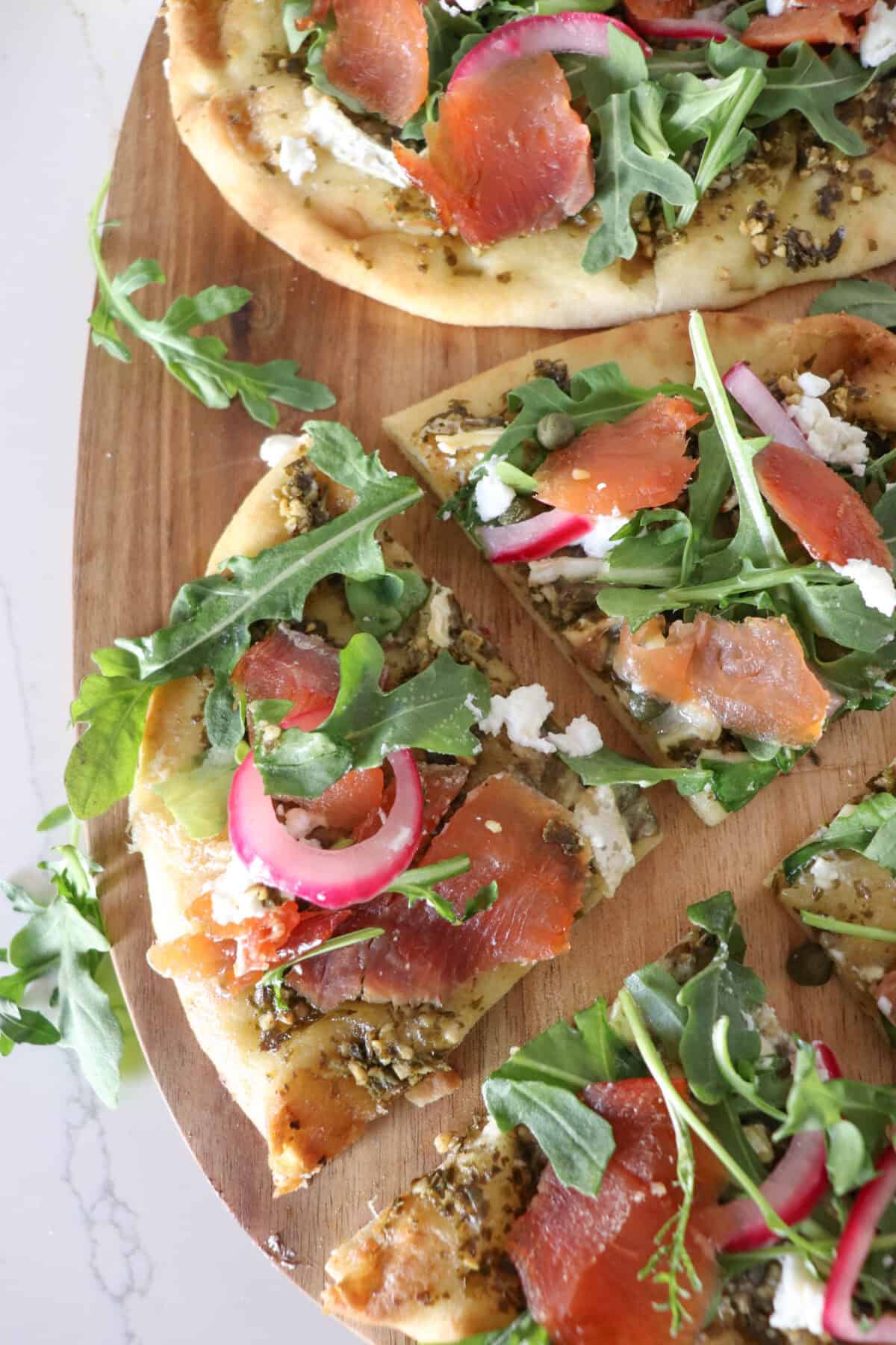 Small round pizza on a wooden cutting board topped with fresh arugula, smoked salmon, goat cheese and pickled onions.