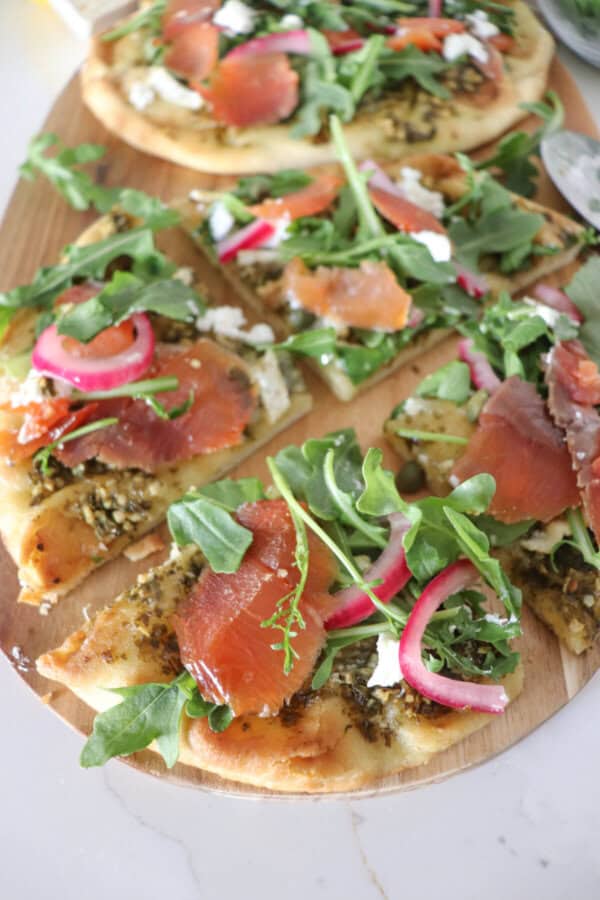 Small round pizza on a wooden cutting board topped with fresh arugula, smoked salmon, goat cheese and pickled onions cut into slices.