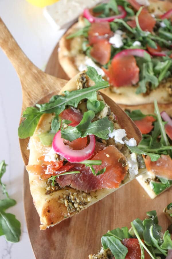 Slice of pizza on a wooden cutting board topped with fresh arugula, smoked salmon, goat cheese and pickled onions.
