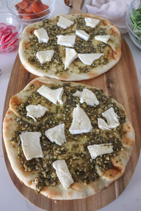 Naan bread covered in pesto sauce with slices of brie on a wooden cutting board.