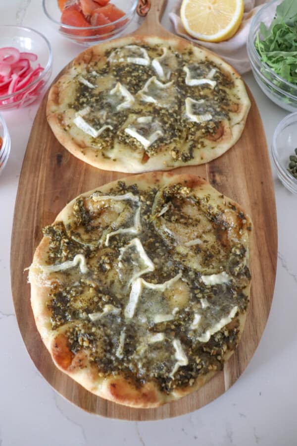 Naan bread covered in pesto sauce with slices of melted brie on a wooden cutting board.