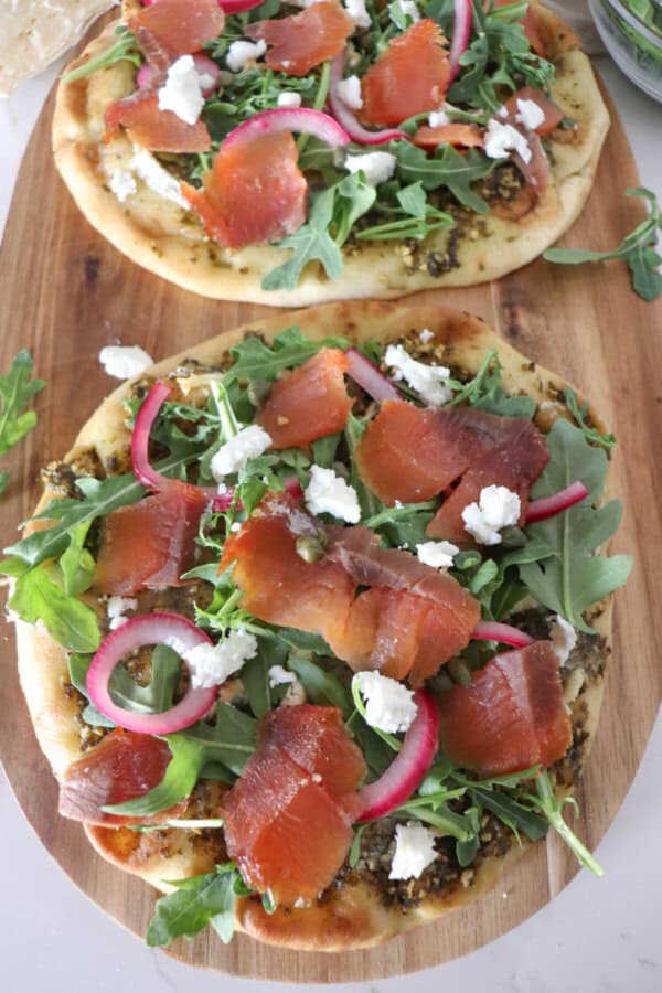 Small round pizza on a wooden cutting board topped with fresh arugula, smoked salmon, goat cheese and pickled onions.