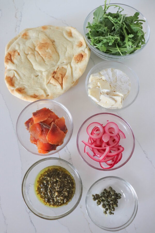 Ingredients for smoked salmon pizza in clear glass bowls on a white counter.