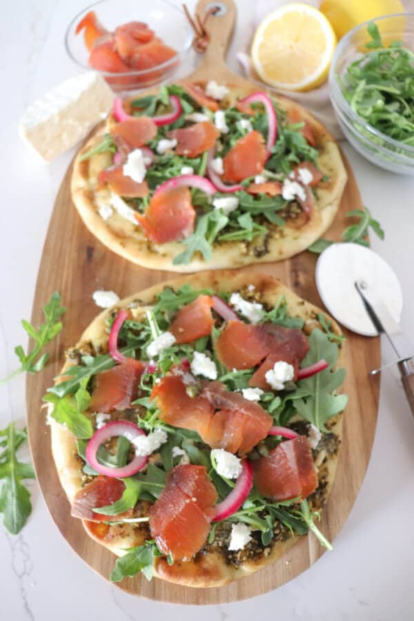 Small round pizza on a wooden cutting board topped with fresh arugula, smoked salmon, goat cheese and pickled onions.