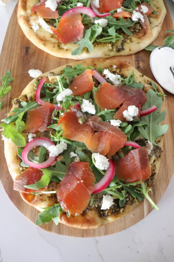 Small round pizza on a wooden cutting board topped with fresh arugula, smoked salmon, goat cheese and pickled onions.