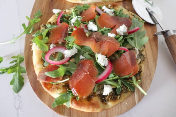 Small round pizza on a wooden cutting board topped with fresh arugula, smoked salmon, goat cheese and pickled onions.