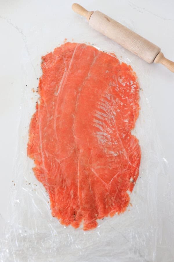 Smoked salmon fillet laid out o plastic wrap on a white counter and rolled flat.