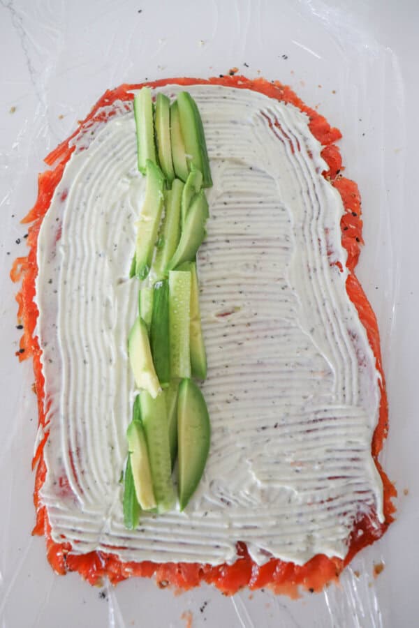 Smoked salmon flattened in a rectangle with cream cheese spread all over it and cucumbers and avocado laid in a row down the center.