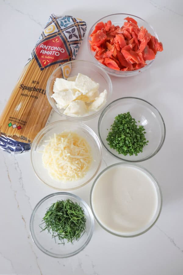 Ingredients for smoked salmon spaghetti in clear glass bowls on a white counter.