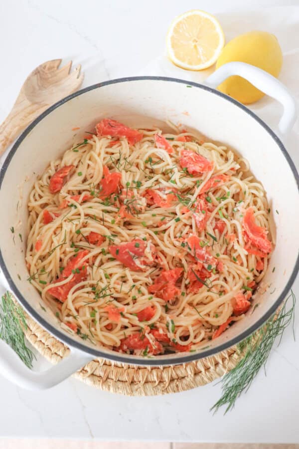 Cooked spaghetti noodles topped with pieces of smoked salmon and garnished with fresh dill in a white dutch oven.
