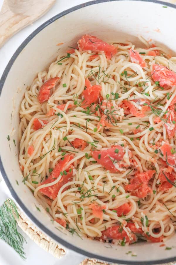 Cooked spaghetti noodles topped with pieces of smoked salmon and garnished with fresh dill in a white dutch oven.