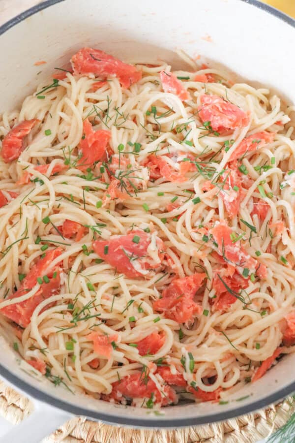 Cooked spaghetti noodles topped with pieces of smoked salmon and garnished with fresh dill in a white dutch oven.