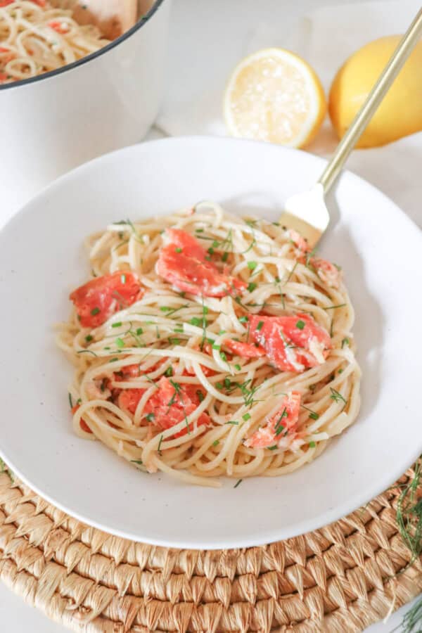 Cooked spaghetti noodles topped with pieces of smoked salmon and garnished with fresh dill in a white bowl.