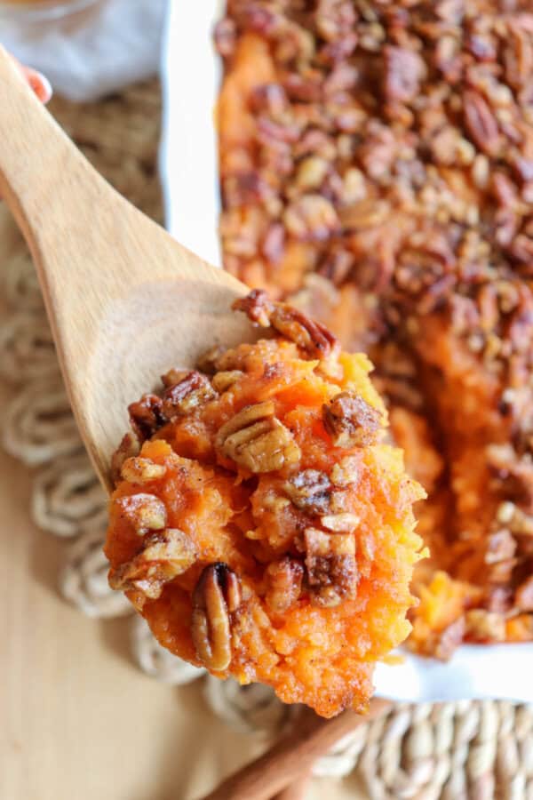 Sweet potato casserole topped with a caramel pecan streusel in a white casserole dish with a scoop taken out and in a wooden spoon.