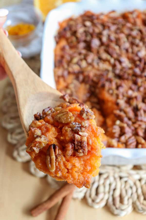 Sweet potato casserole topped with a caramel pecan streusel in a white casserole dish with a scoop taken out in a wooden spoon.