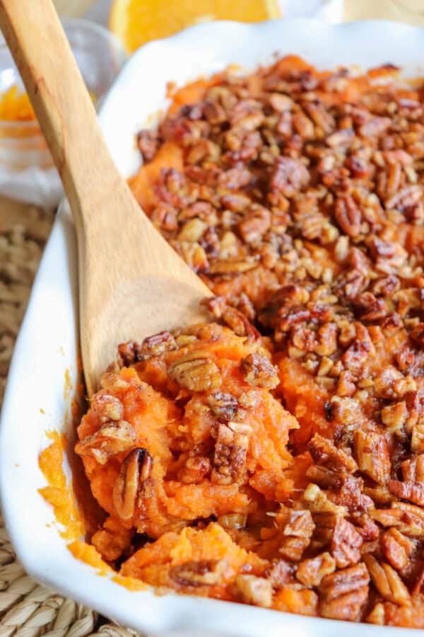 Sweet potato casserole topped with a caramel pecan streusel in a white casserole dish with a wooden spoon scooping out some of the mixture.