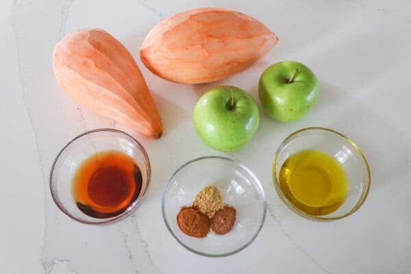 Ingredients for sweet potato and apple sheet pan dinner in clear glass bowls on a white counter.