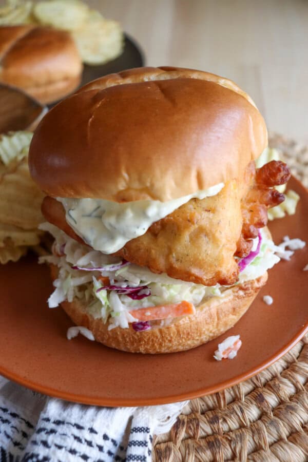 Fried fish filet on a brioche bun with coleslaw and tartar sauce on an orange plate.
