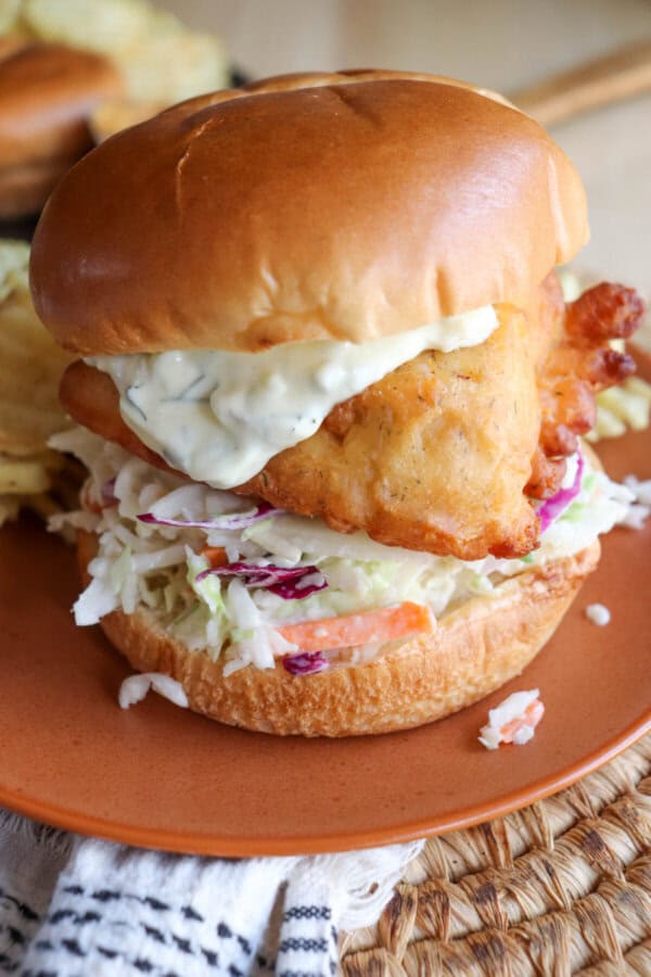 Fried fish filet on a brioche bun with coleslaw and tartar sauce on an orange plate.