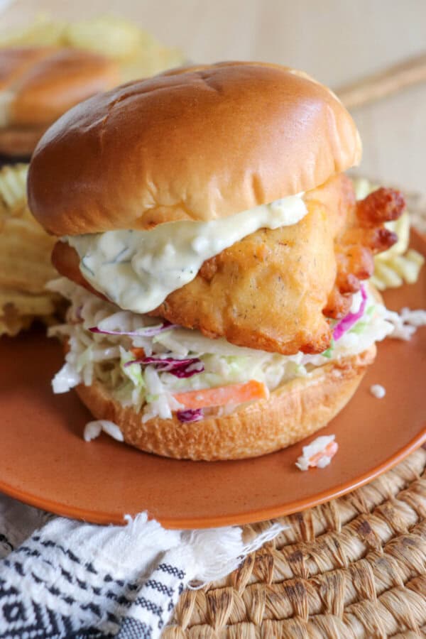 Fried fish filet on a brioche bun with coleslaw and tartar sauce on an orange plate.