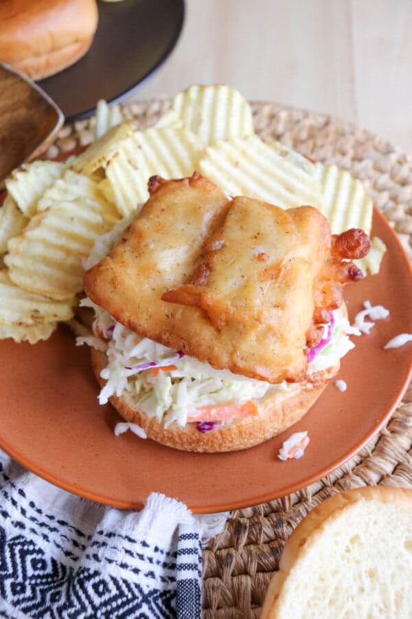 Fried fish filet on a brioche bun with coleslaw and tartar sauce on an orange plate.