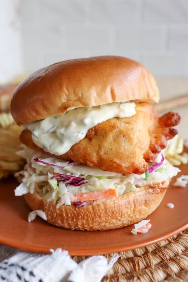 Fried fish filet on a brioche bun with coleslaw and tartar sauce on an orange plate.