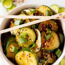 Sliced cucumbers in chili dressing sprinkled with sesame seeds and diced green onions in a white bowl.