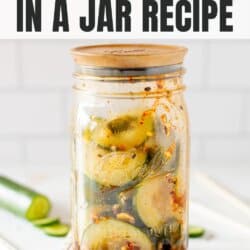 Sliced cucumbers, chili dressing and sesame seeds in a clear glass jar on a white counter with a white tile background.