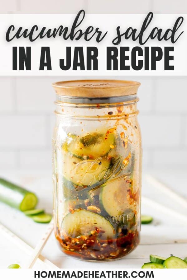 Sliced cucumbers, chili dressing and sesame seeds in a clear glass jar on a white counter with a white tile background.