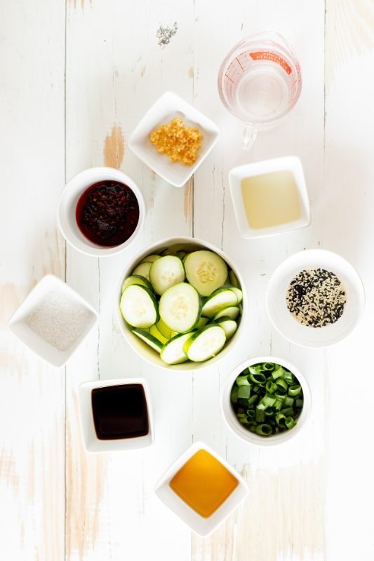 Ingredients for cucumber salad in small bowls on a white table.