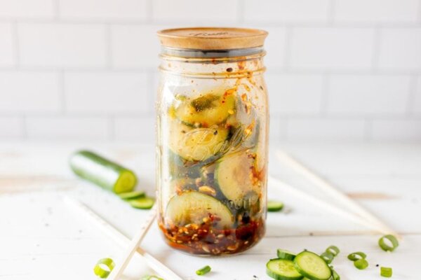 Sliced cucumbers, chili dressing and sesame seeds in a clear glass jar on a white counter with a white tile background.