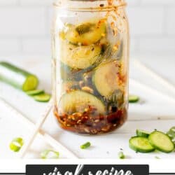 Sliced cucumbers, chili dressing and sesame seeds in a clear glass jar on a white counter with a white tile background.