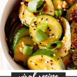 Sliced cucumbers in chili dressing sprinkled with sesame seeds and diced green onions in a white bowl.
