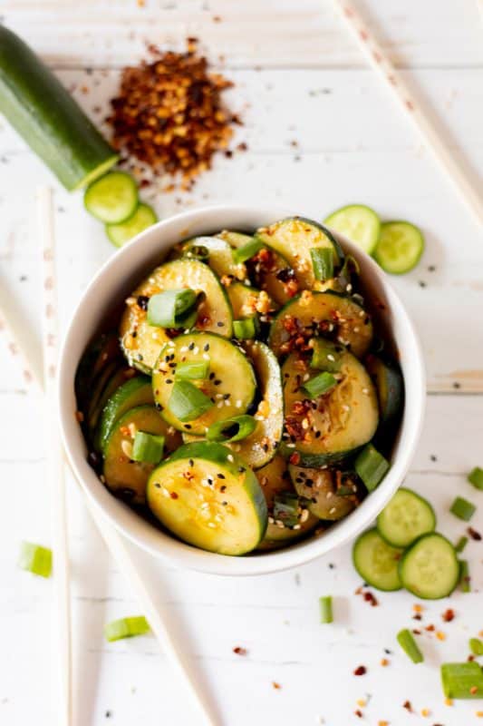 Sliced cucumbers in chili dressing sprinkled with sesame seeds and diced green onions in a white bowl.
