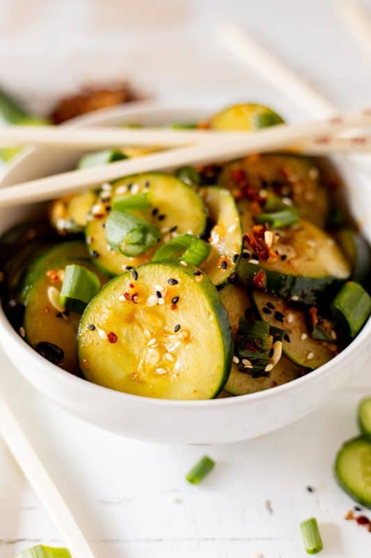 Sliced cucumbers in chili dressing sprinkled with sesame seeds and diced green onions in a white bowl with wooden chopsticks.