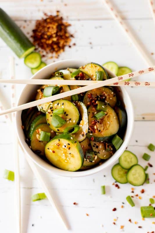 Sliced cucumbers in chili dressing sprinkled with sesame seeds and diced green onions in a white bowl with wooden chopsticks.