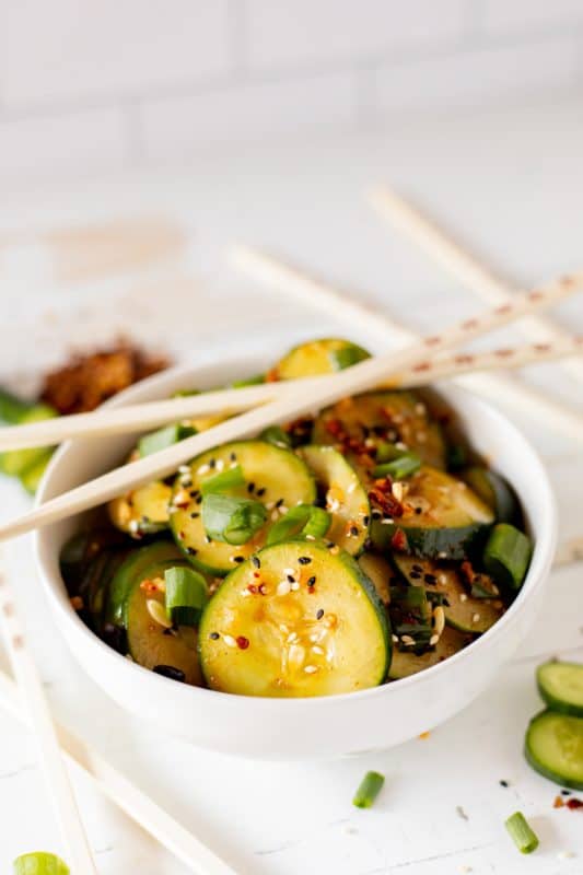 Sliced cucumbers in chili dressing sprinkled with sesame seeds and diced green onions in a white bowl with wooden chopsticks.