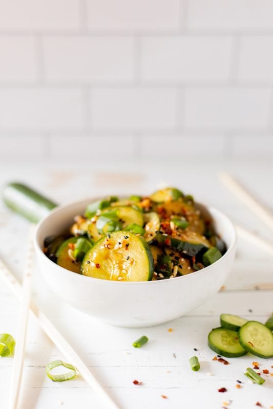 Sliced cucumbers in chili dressing sprinkled with sesame seeds and diced green onions in a white bowl.