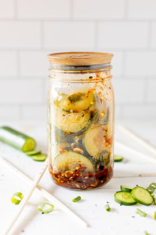 Sliced cucumbers, chili dressing and sesame seeds in a clear glass jar on a white counter with a white tile background.